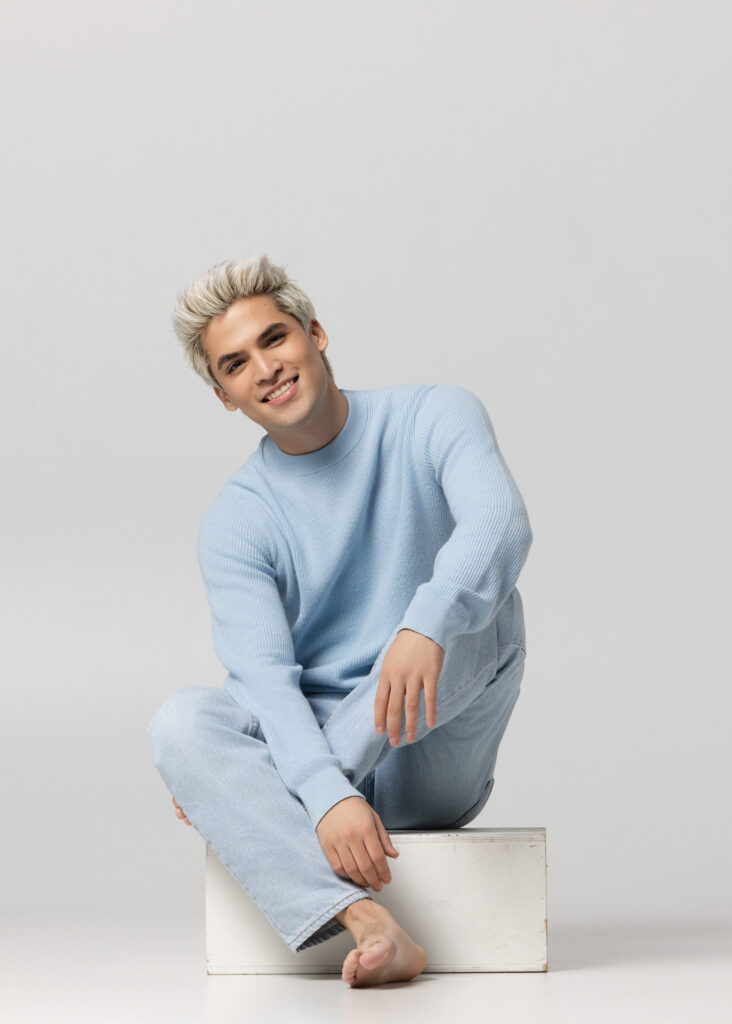 Male model in a light blue knit sweater and denim seated on a white studio block, model portfolio by Ashkan Image, Houston