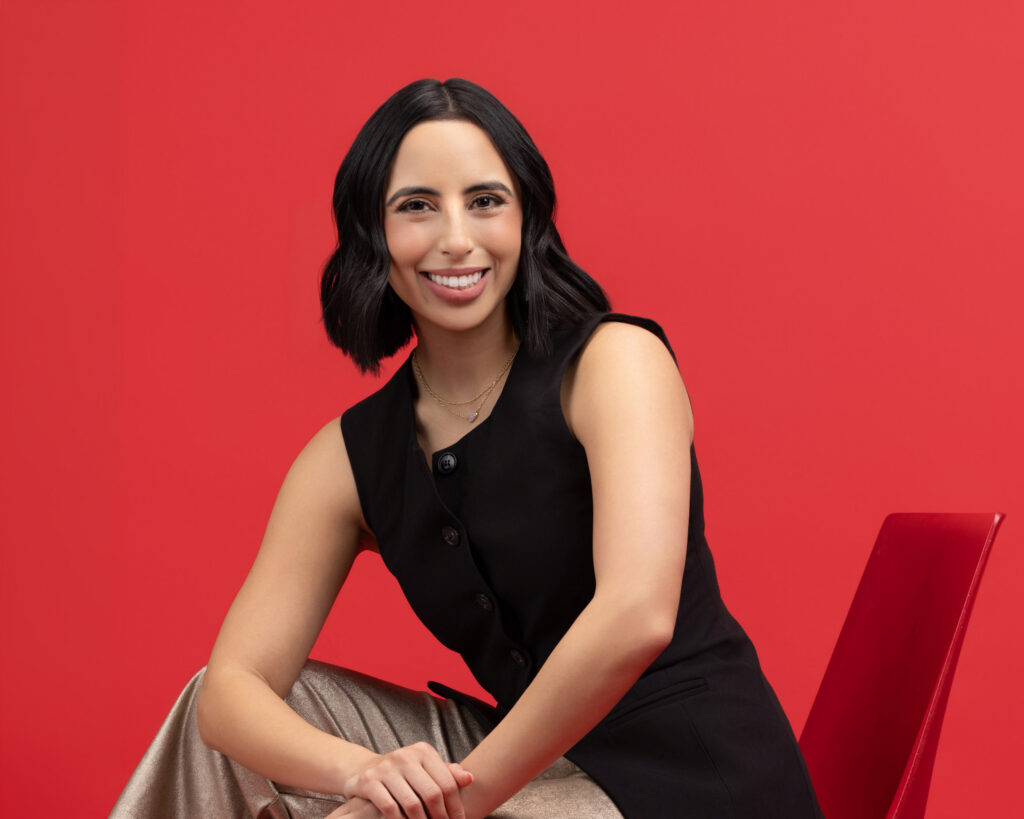 Personal branding photography in Houston, woman in a black vest seated and smiling against a red backdrop, by Ashkan Image